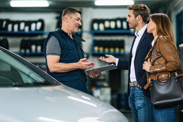 Young happy couple talking to car mechanic in auto repair shop.