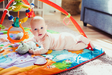 4 months old baby girl lying on colorful play mat on the floor. Activity carpet for kids. Early development at home.