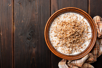 Buckwheat porridge with milk in ceramic bowl on dark wooden background. Top view, copy space.