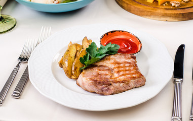 Grilled pork with potatoes on a white plate with hot snack, salads, cutlery, wine and water glasses on banquet restaurant table background. European food in a restaurant setting.