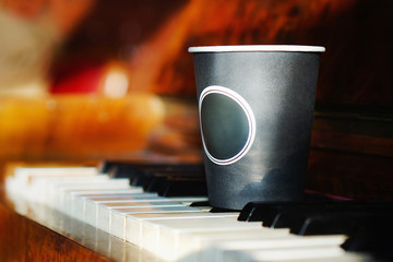 Black paper cup of coffee or tea on the piano keys, break during music lessons. Round sticker with copy space on the cup. Place for text