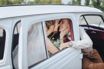 Wedding closeup portrait of smiling newlyweds in car window. Stylish groom in a suit and a beautiful bride in a white dress are sitting on a car armchair. Photography and concept.