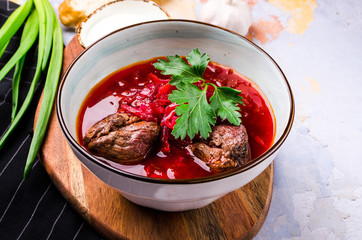 Traditional Ukrainian Russian Borscht with bacon, hot soup with beets, cabbage and carrots in a bowl on a wooden board. Served with green onions, sour cream, rye bread, garlic, greenery.