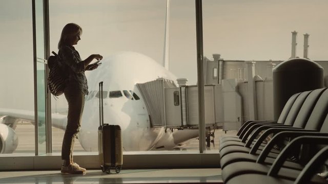 A woman with a passport and a boarding pass is standing at a large window at the airport, outside the window is a huge airliner. Start of the journey