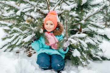 Happy little girl in winter clothes sitting outdoor under spruce tree covered with snow