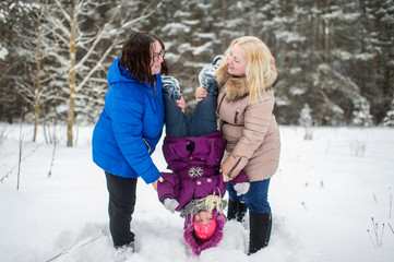 Two adult positive woman holding little girl legs upside down in winter snowy field.
