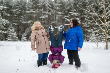 Two adult positive woman holding little girl legs upside down in winter snowy field.