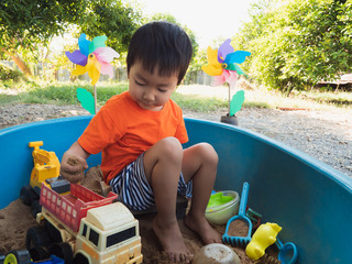 Asian child boy in orange t-shirt playing toy and sand in sandbox with happy face outdoor with rural natural background. Family relax and freedom time in summer holiday.