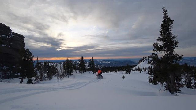travel snowmobile, in a beautiful mountain valley at sunset with colorful sky and rocks in the forest. on the background of the winter forest, leaving behind a trail of splashes of white snow. 4K