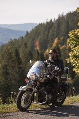 Handsome bearded biker in black leather jacket and sunglasses resting on cruiser motorcycle on country roadside, on blurred background of foggy green hills covered with dense spruce forest.