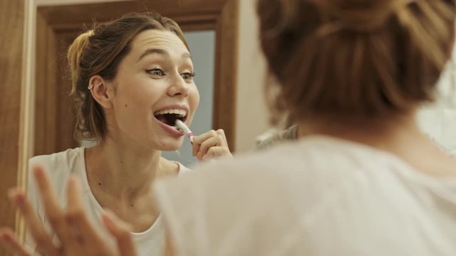 An attractive smiling woman looking to the mirror while cleaning her teeth in the bathroom with brush and toothpaste