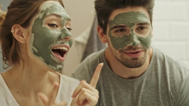 Happy positive young couple man and woman having fun while standing in a bathroom with green face mask