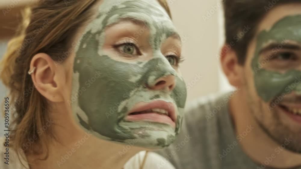 Poster Close up view of an attractive young woman looking at herself while standing in the bathroom with a green mask on her face