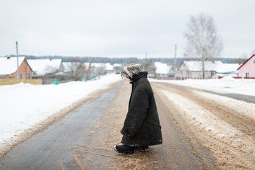 Strange little girl in worn oversized clothes standing at snowy road win winter day in russian village.