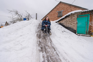 Cheerful girl using plastic bag as a sled and riding down from cellar with icey trek in front of brick village house in winter day.