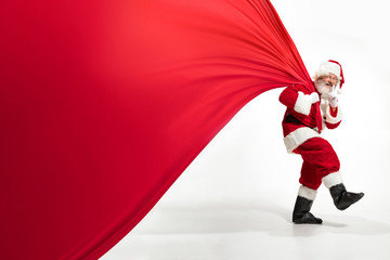 Santa Claus pulling huge bag full of christmas presents isolated on white background. Caucasian male model in traditional costume. New Year 2020, gifts, holidays, winter mood. Copyspace for your ad.