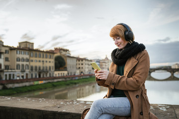 Fototapeta na wymiar Young woman with red hair listens to music with headphones and uses the smartphone sitting on a bridge in Florence - Millennial is having fun and takes some time off