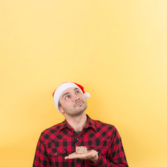Pensive man in a red hat, holds a gift with interest on his face. Positive human emotions for christmas, secret santa. Yellow background, copy space.