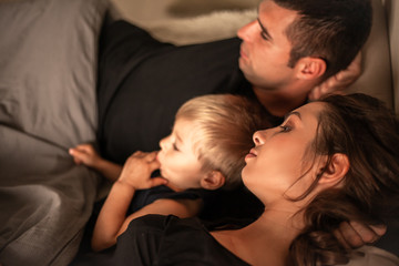 Happy family at home bedroom lies with kid on the bed together under the blanket. People wearing black t-shirt,  shorts and baby bodysuit.