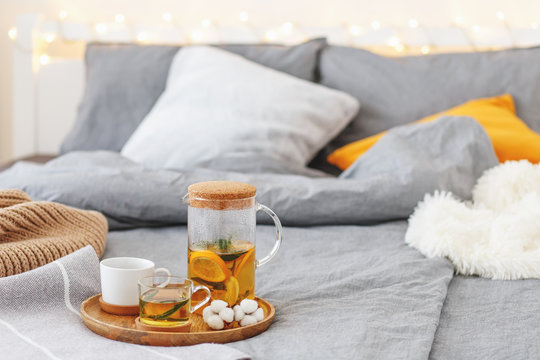 Cozy Winter Home Decor: Glass Teapot And Two Cups Of Citrus Tea With Orange, Lemon And Mint On A Wooden Tray, Bed With Grey Linen, Blanket, Pillows, Plaid, Led Garland Light. Breakfast In Bed.