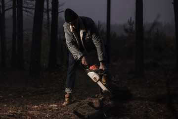 Obraz premium Lumberman work with a chainsaw in the forest. Strong lumberjack with the chainsow in the forest.Stylish lumberman getting ready for work. Lifestyle. Hard work in the forest. Illegal logging.
