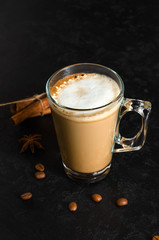 Transparent mug of Cappuccino or latte, some scattered coffee beans, cinnamon sticks, anise star on black background. Morning hot beverage. Closeup. Copy space