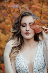 Autumn mood! happy smiling woman holding in her hands yellow maple leaves hiding her eye on the background of orange leaves. Brunette with curly hair and make up. Female fashion.