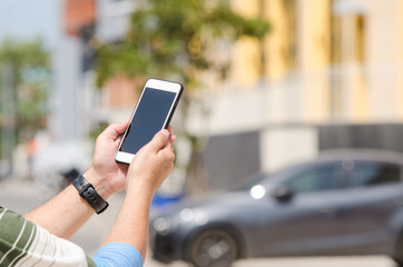 Hands are holding touch screen smart phone on blurred building and car background.