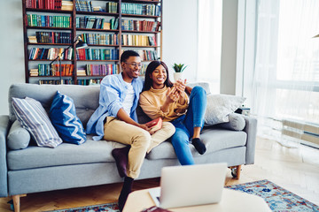 Cheerful African American couple having fun together at cozy living room sitting on sofa with smartphone, young hipster guys taking selfie on mobile phone camera recreating on leisure at home interior