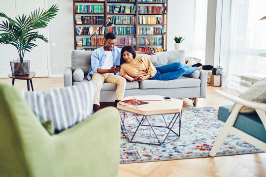 Positive African American Couple In Love Resting Together At Apartment On Comfortable Sofa, Dark Skinned Male Showing New App On Smartphone To His Girlfriend Lying With Book In Modern Home Interior