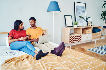 African american couple in love talking to each other sitting at bed room using laptop computer and smartphone for networking, dark skinned young family having conversation resting on free time.
