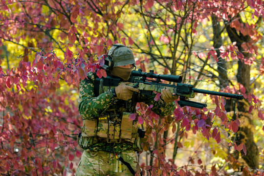 Closeup man in green uniform with sniper rifle. Airsoft soldier stand in red bush. Horizontal photo