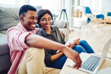 Fototapeta premium Cheerful african american couple satisfied with resting together in cozy interior living room watching movie on laptop computer, smiling dark skinned male and female laughing at funny videos.
