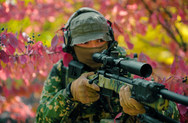 Closeup man in green uniform with sniper rifle. Airsoft soldier stand in red bush