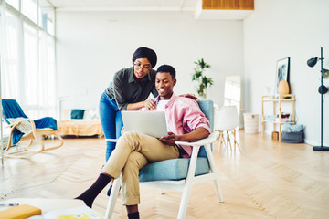 Positive african american romantic couple making shopping together at home interior using laptop computer, smiling dark skinned woman looking at her husband work online on freelance via netbook .