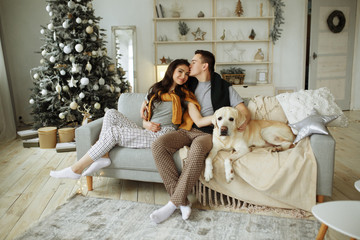 happy couple is sitting near sofa with a dog, christmas. new year at home. family and date