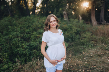 A young and beautiful pregnant blonde girl in a white dress stands and holds on to her stomach at sunset. Portrait of a pretty woman expecting a baby in nature. Pregnancy picture, concept.