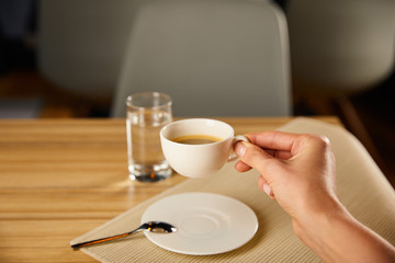 cropped view of woman holding cup of coffee near glass of water in cafe