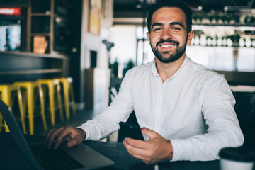 Portrait of cheerful male entrepreneur in casual white shirt holding cellphone device and smiling at camera while doing distance job on modern laptop computer, happy man using 4g on digital technology