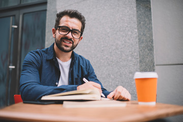 Fototapeta na wymiar Portrait of clever male student in casual apparel and classic glasses for vision correction sitting at street cafeteria and looking at camera during time for learning and preparing to university exams