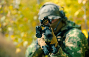 Closeup airsoft man in uniform, stand with sniper rifle on yellow forest backdrop. Soldier aims at...
