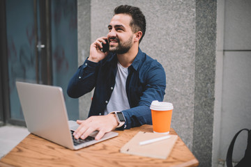 Happy male programmer in casual wear enjoying break for smartphone calling to best friend using roaming connection outdoors, cheerful man making international conversation via mobile gadget