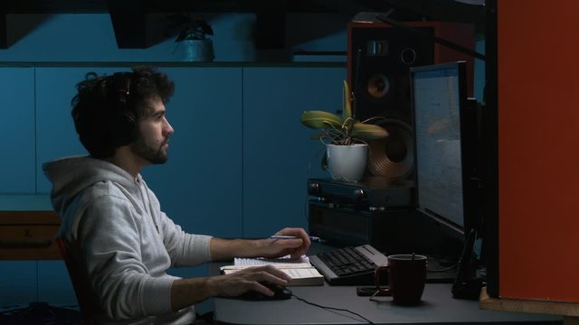 Young adult office worker sitting at desk in modern workspace late at night / active student working at computer at evening in dark room, blue background moonlight.