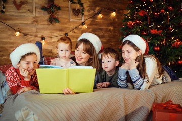 Mother reads a book with children lying on the bed in the room with tree at Christmas.