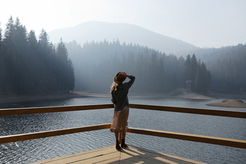 happy young woman in hat enjoys lake view in mountains . Relaxing moments in forest. Back view of...