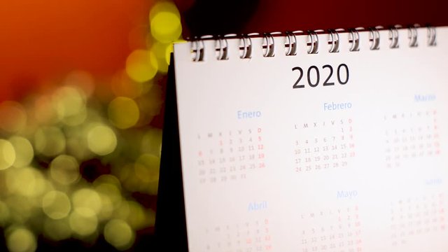 Semi-lateral view of a white calendar of the new year 2020 with bokeh effect in the background and flickering colored lights that turn on and off. New Year calendar with Christmas lights background