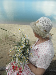 Beautiful blond woman in a hat and white with a pink pattern dress with a bouquet of daisies on the shore of the pond. Side view / view from above