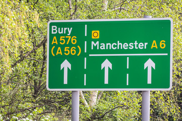 Green road signs on a leafy background showing directions to manchester and bury including the A6 A576 and A56.