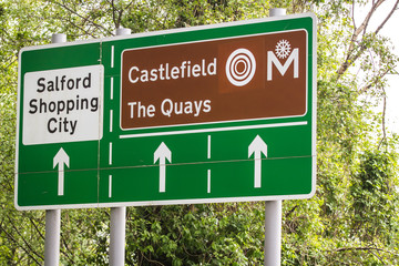 Green road signs on a leafy background showing directions to salford shopping city and and castlefield in greater manchester.