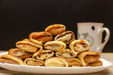 Homemade pancakes with filling stacked in a pyramid on a white plate against a black wall with a Cup, copy space